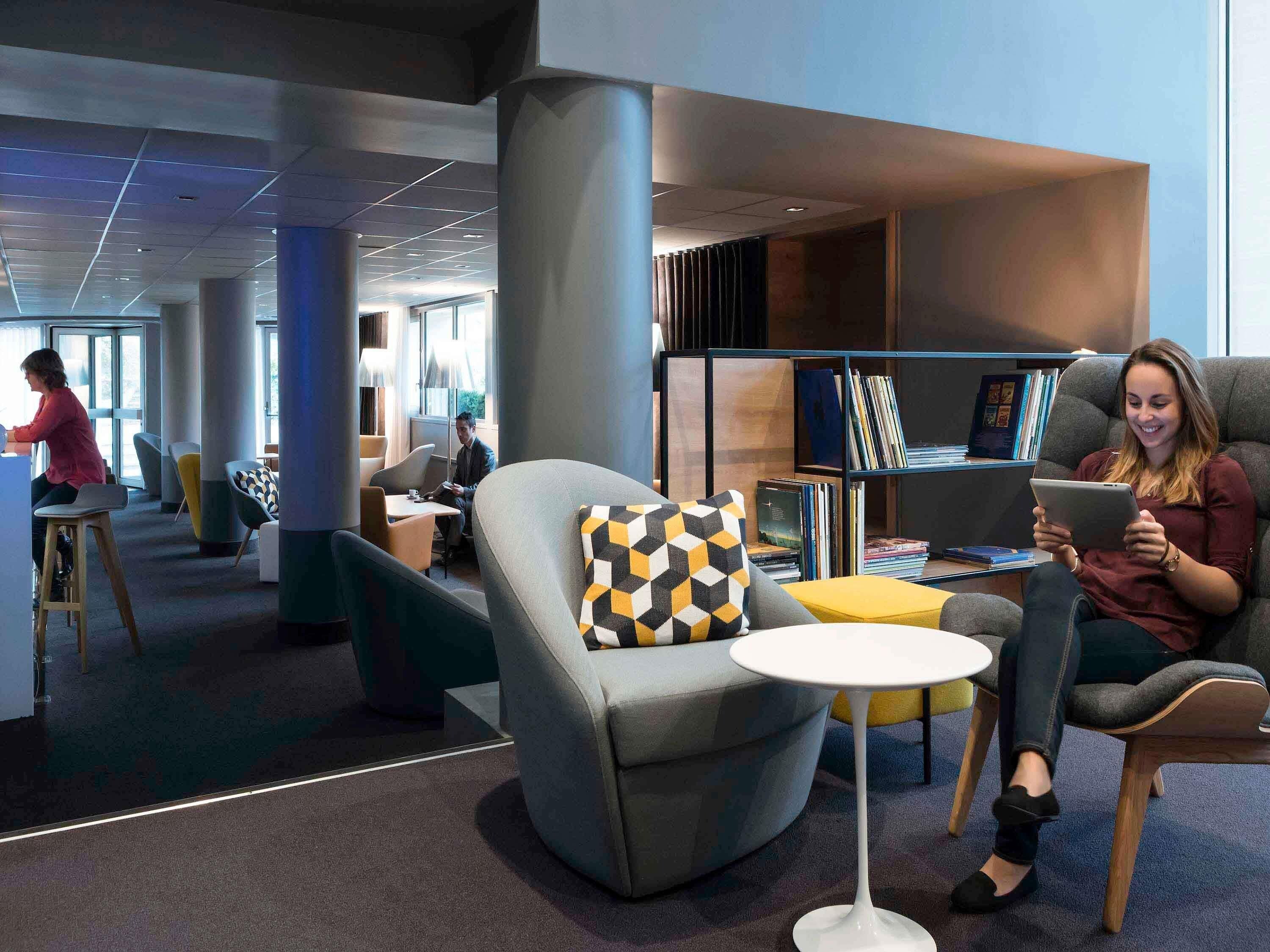 A modern library with seating areas, bookshelves, and a person using a tablet.