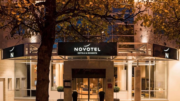 A hotel entrance with a sign reading 'NOVOTEL HOTELS & RESORTS,' a glass facade, and a tree in the foreground.