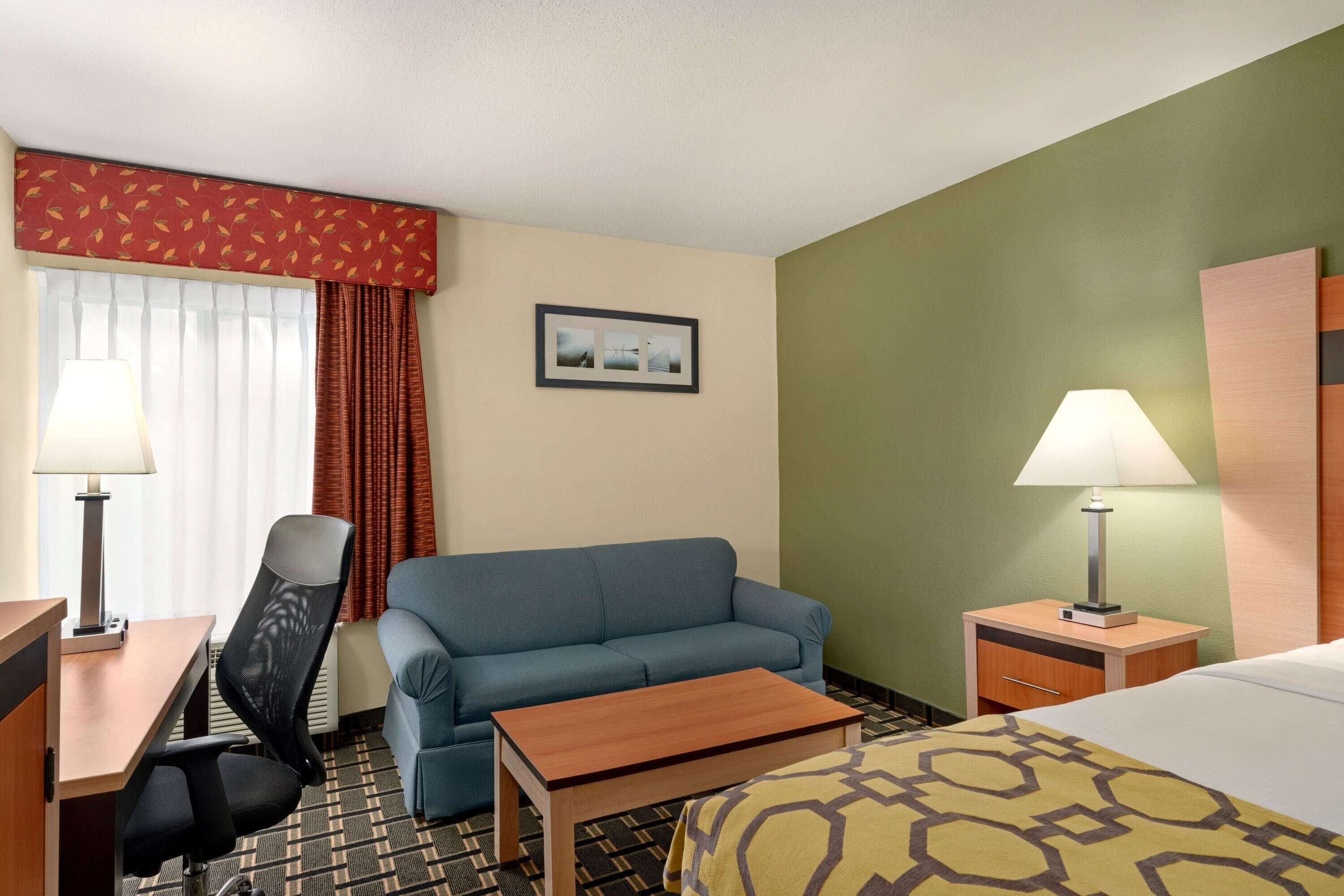 A hotel room with a blue sofa, a wooden table, a desk with a lamp, a bed with a patterned blanket, a nightstand with a lamp, and a picture on the wall.