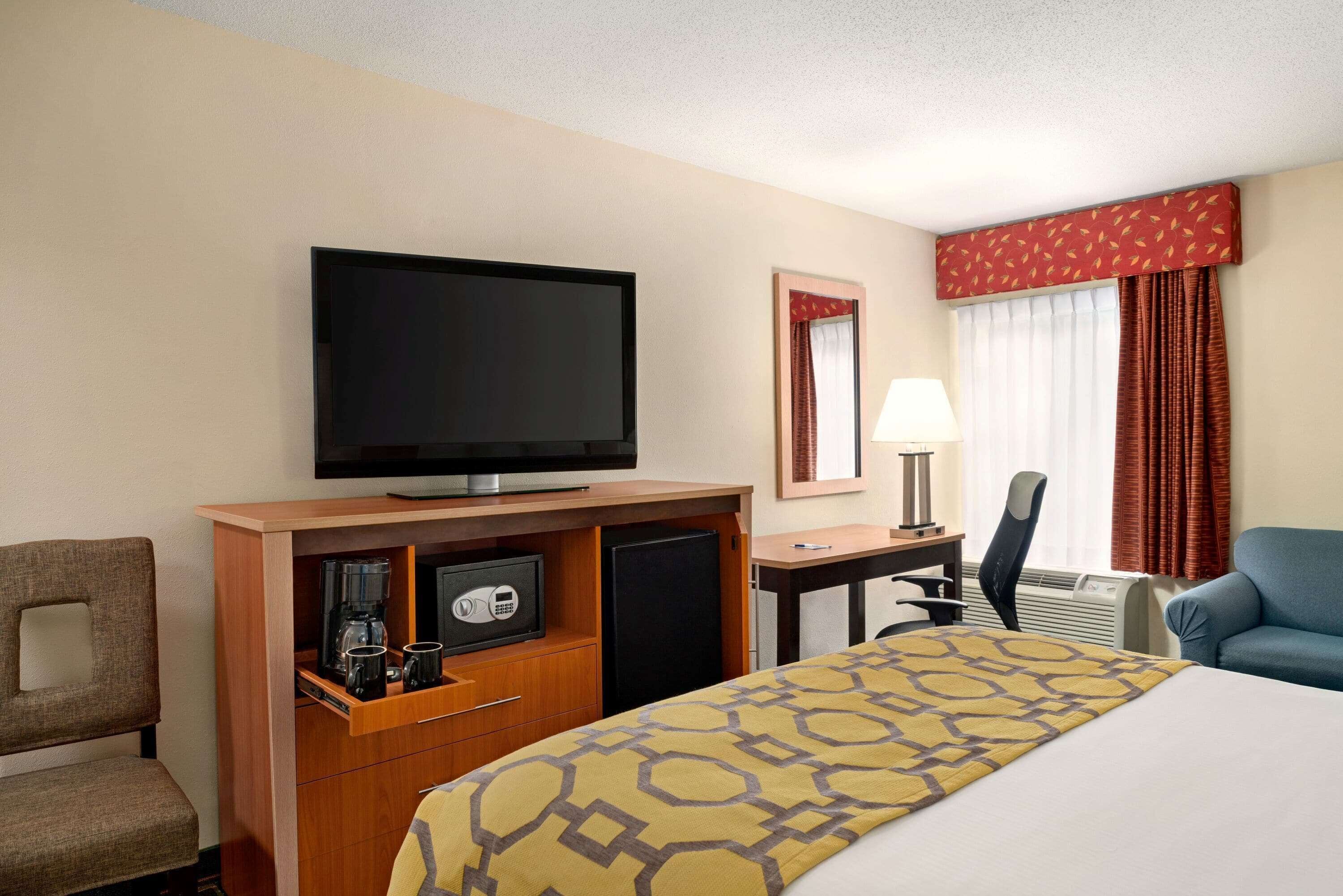 A hotel room with a bed, a television, a desk with a lamp, a chair, and a safe.