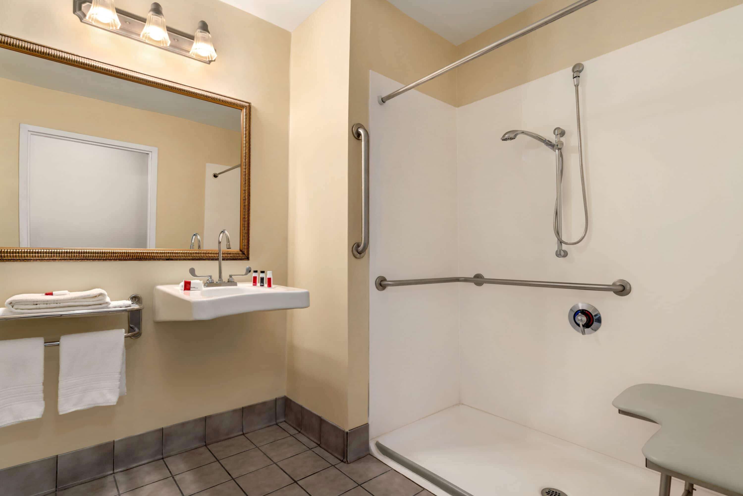 A bathroom with a sink, mirror, towel rack, shower with grab bars, and a bench.