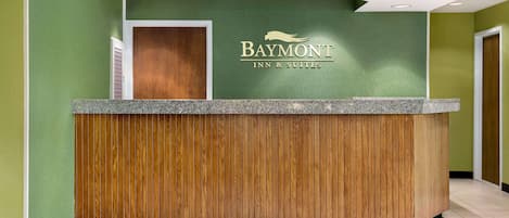 Un mostrador de recepción con diseño de panel de madera, una pared verde con el logotipo de Baymont Inn & Suites y un techo blanco con iluminación empotrada.