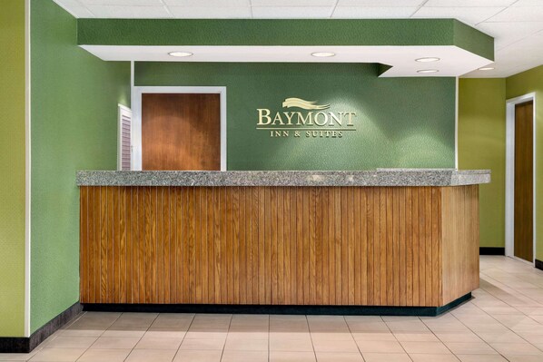 A reception desk with a wooden panel design, a green wall with the Baymont Inn & Suites logo, and a white ceiling with recessed lighting.