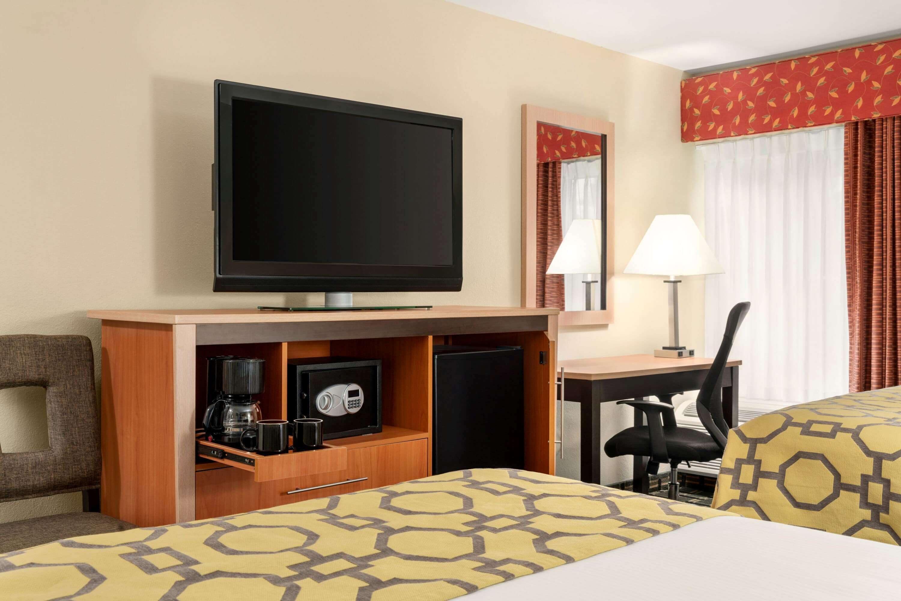 A hotel room with a bed, a desk, a chair, a television, a coffee maker, a safe, and a mirror.