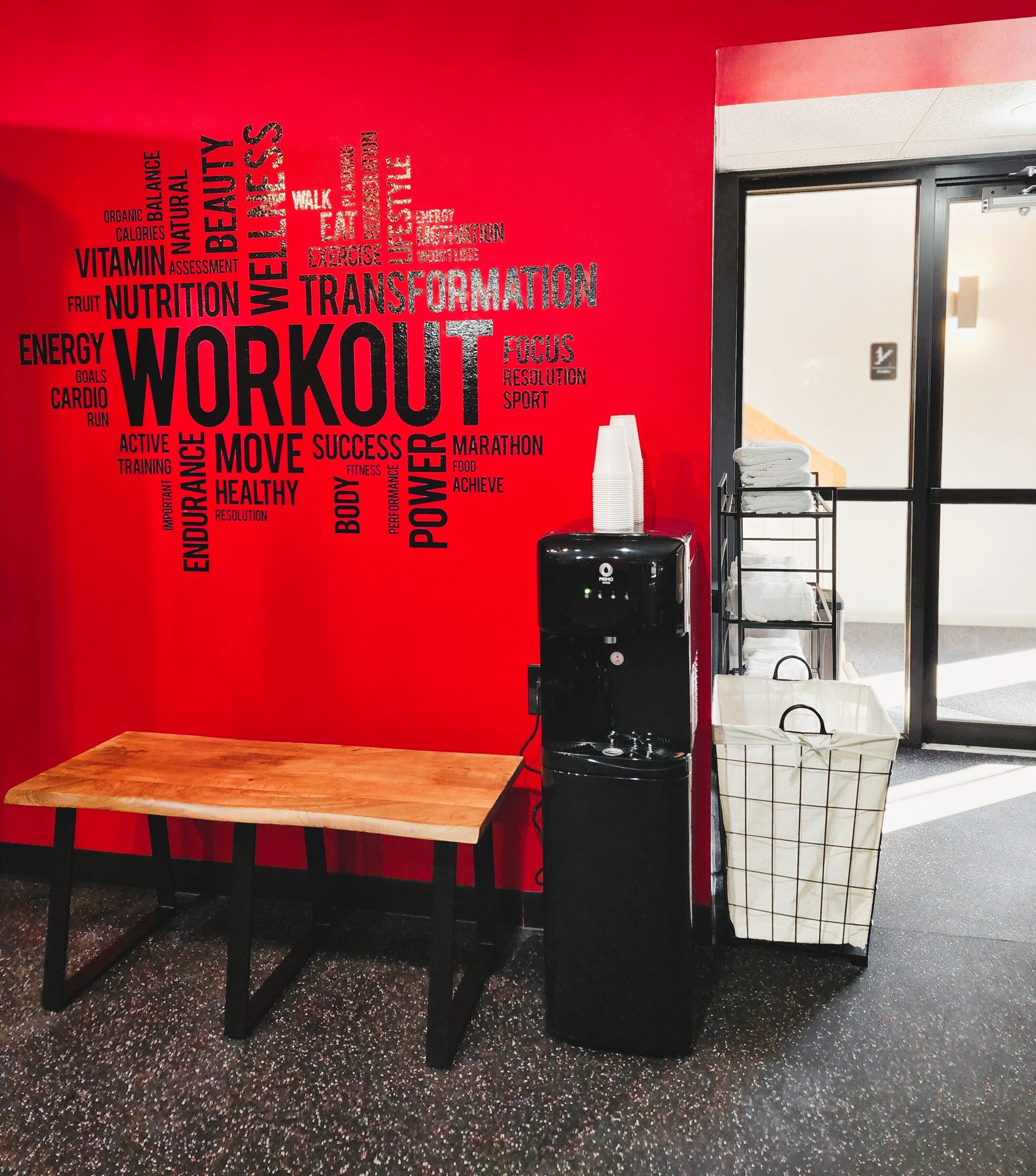 A red wall with a word cloud related to fitness and health, a wooden bench, and a black appliance, likely a coffee machine, in a commercial setting.