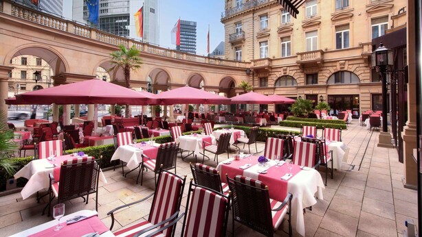 Ein Straßencafé mit Tischen und Stühlen, umgeben von einem historischen Gebäude mit Arkaden und einem modernen Wolkenkratzer im Hintergrund.