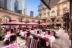 Un café al aire libre con mesas y sillas, rodeado por un edificio histórico con arcos y un rascacielos moderno al fondo.