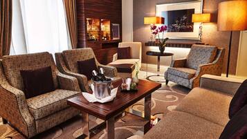 A modern hotel lobby with a seating area featuring two armchairs and a sofa, a wooden coffee table with a bottle of champagne, and a small side table with a vase of flowers.