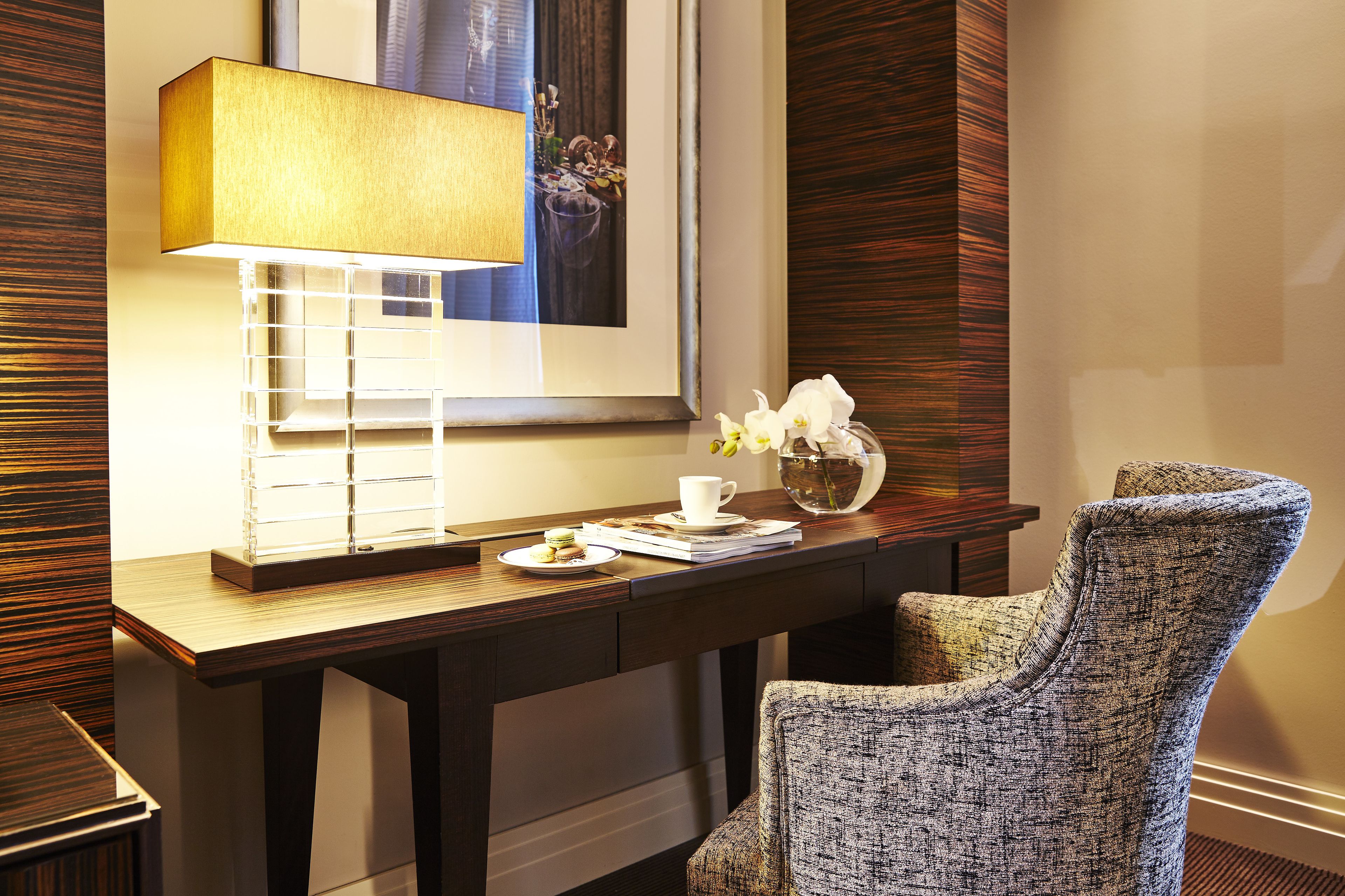A hotel room with a wooden desk, a lamp, a framed picture, a chair, and a small table with a cup and flowers.