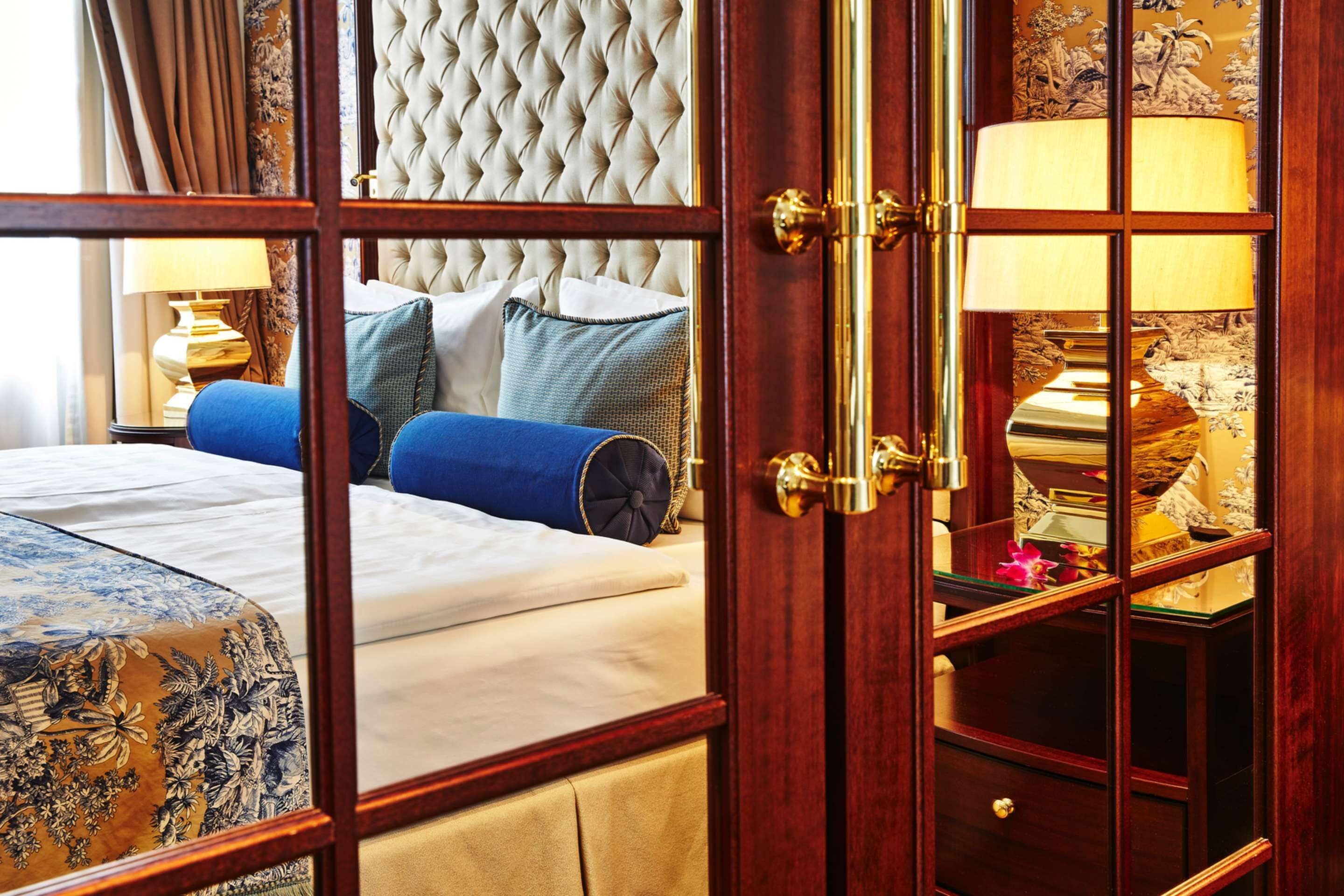 Habitación de hotel con un cabecero capitonado, una cama con ropa de cama azul y gris, una mesita de noche de madera con una lámpara y una mesa con estampado floral.