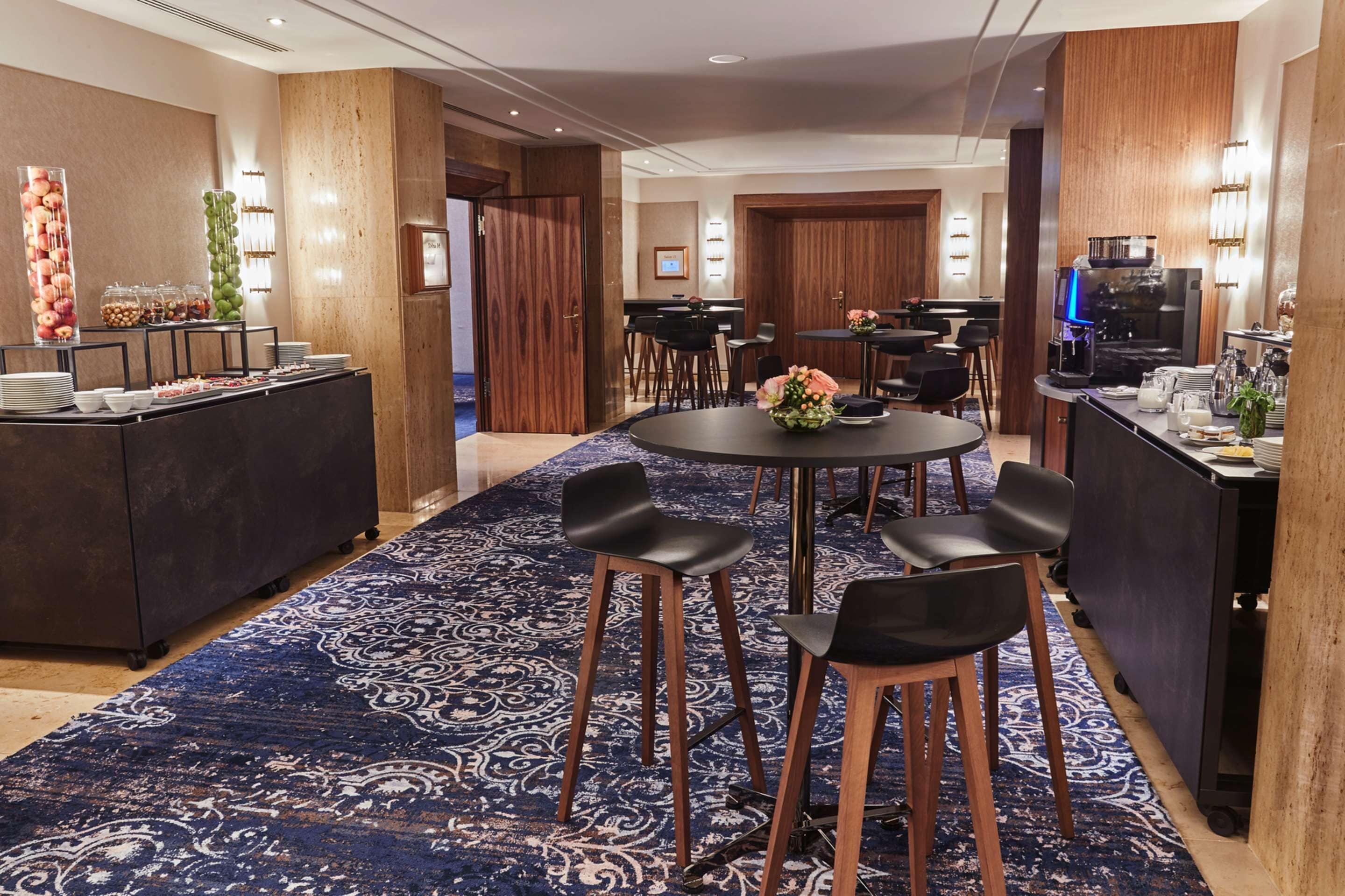 Ein Hotelzimmer mit einem runden Esstisch, schwarzen Stühlen, einer Büfettstation mit Obst und Snacks sowie einer Küchenzeile mit einer Kaffeemaschine und einem großen, gemusterten Teppich.