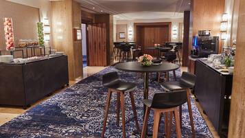 Ein Hotelzimmer mit einem runden Esstisch, schwarzen Stühlen, einer Büfettstation mit Obst und Snacks sowie einer Küchenzeile mit einer Kaffeemaschine und einem großen, gemusterten Teppich.