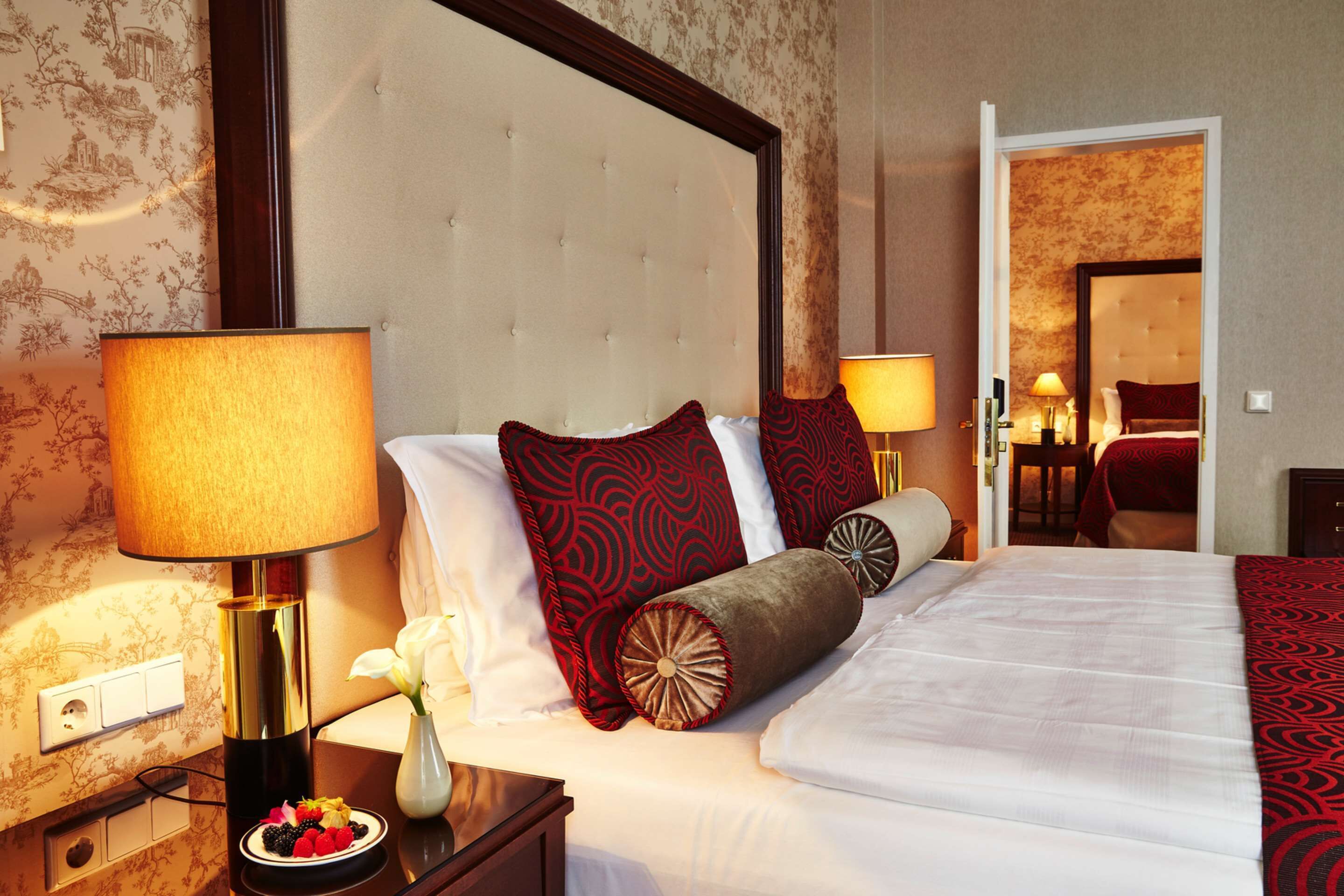 Ein Hotelzimmer mit einem großen Bett, einem Nachttisch mit Lampe, einer Vase mit Blumen und einer Schale mit Obst.