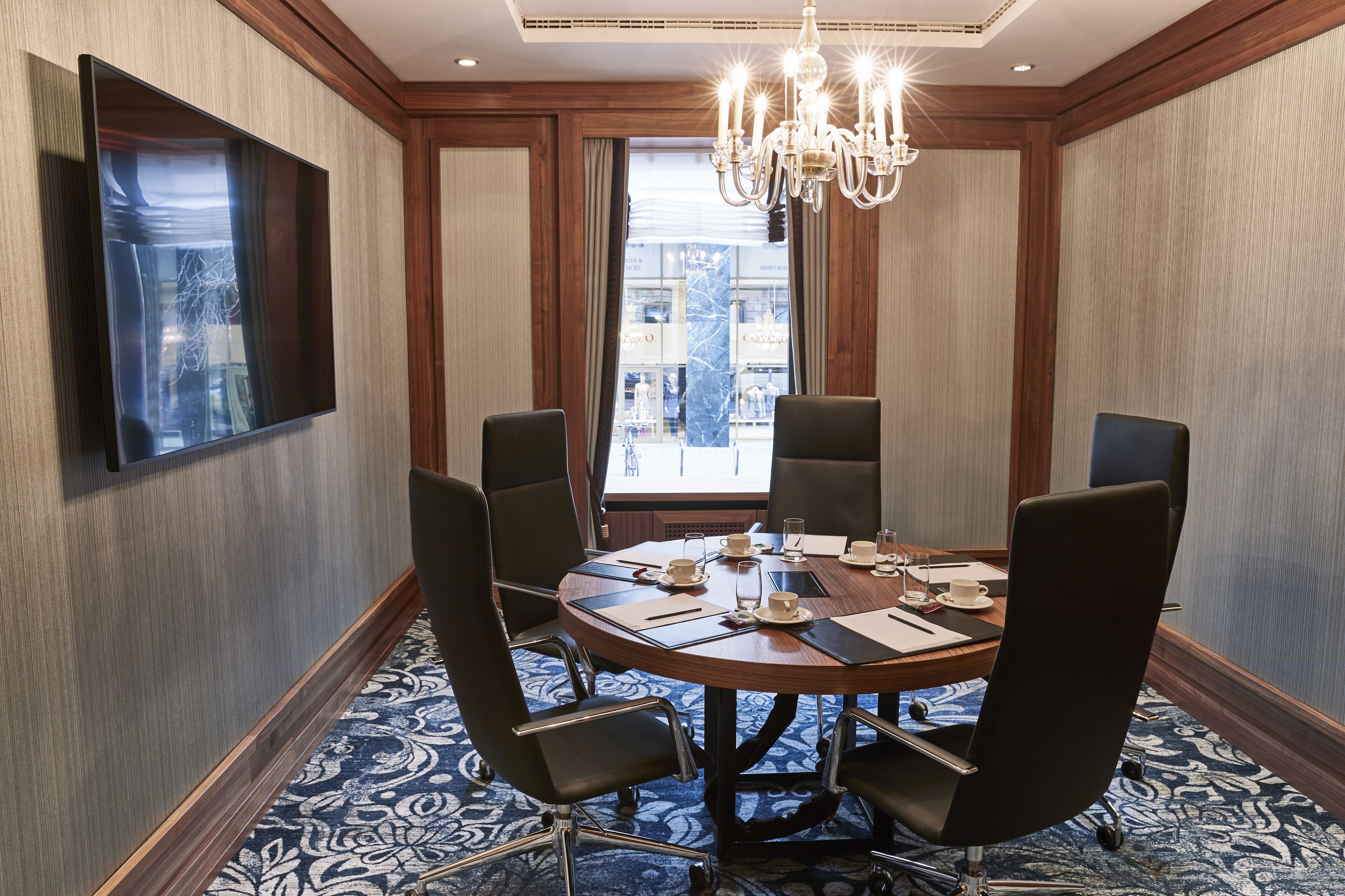 A conference room with a round table, six chairs, a chandelier, and a large window with a view of a building.