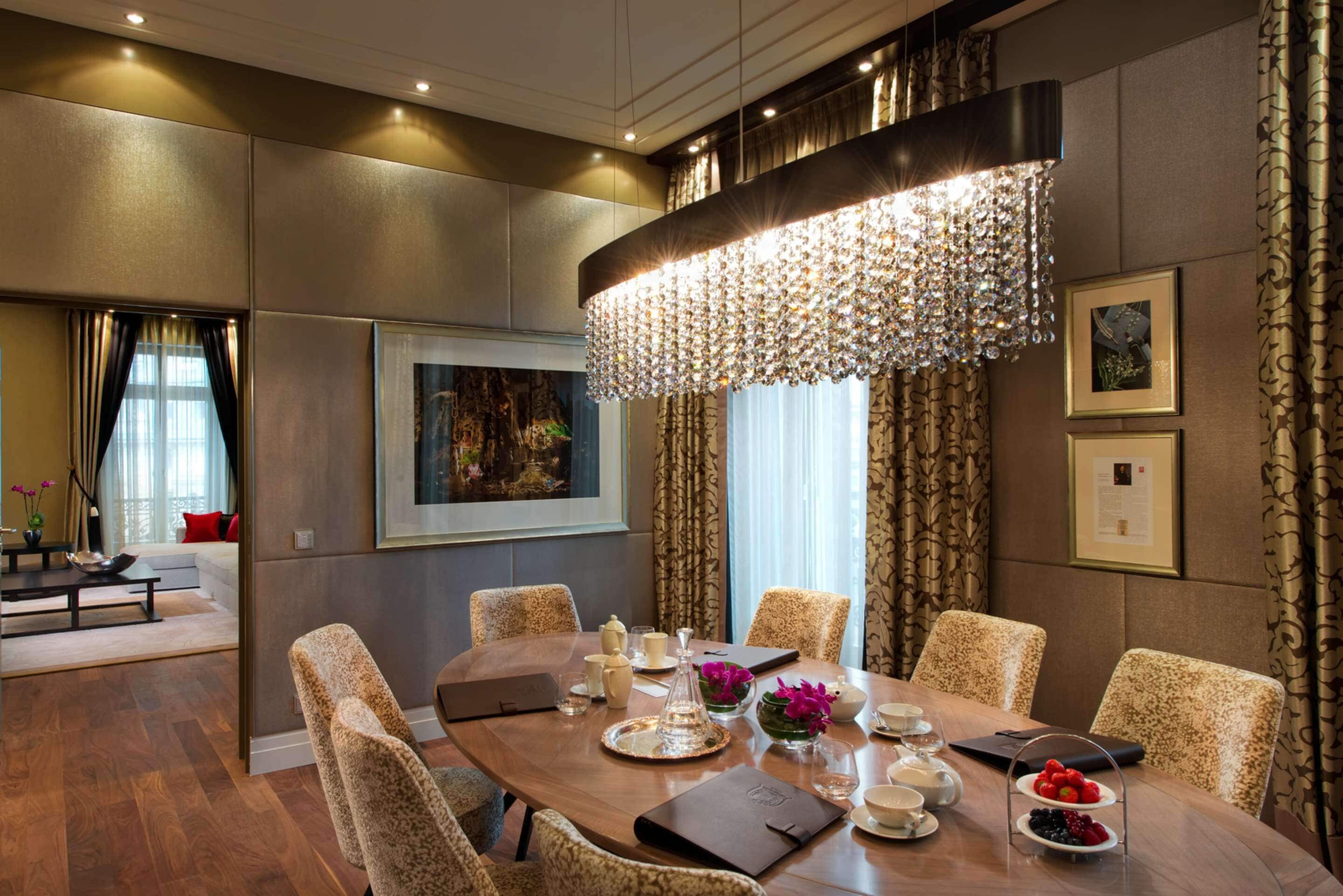 A dining area with a large table, chairs, and a chandelier. There are framed pictures on the walls and a sofa in the background.