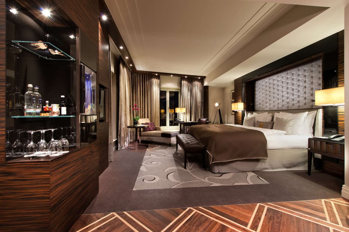 Ein Hotelzimmer mit einem großen Bett, einem Nachttisch, einer Sitzecke mit Sofa und Sessel, einer Vitrine mit Flaschen und einem gemusterten Teppich.
