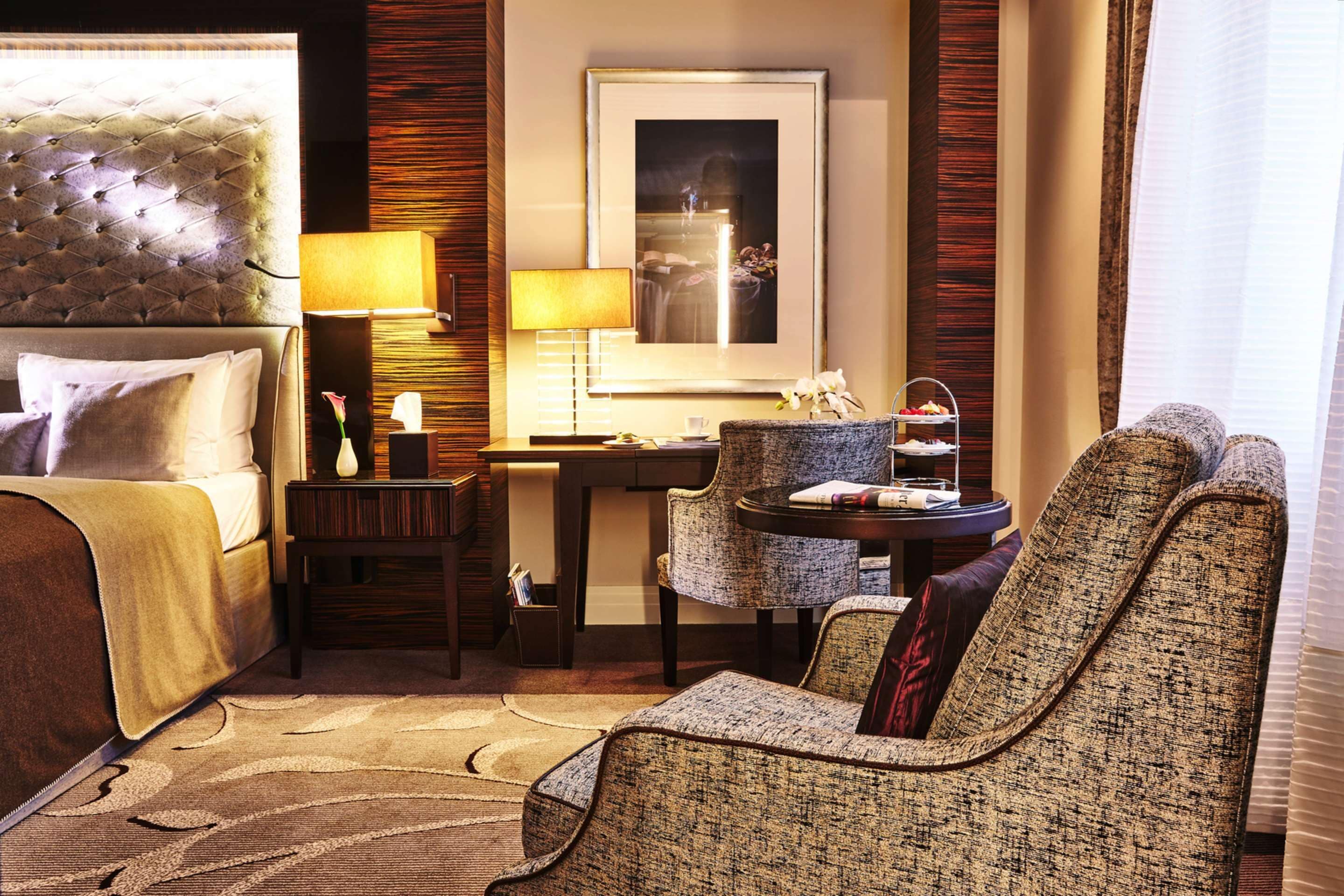A hotel room with a tufted headboard, a bed with pillows, a desk with a lamp, a chair, and a framed picture on the wall.