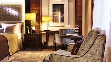A hotel room with a tufted headboard, a bed with pillows, a desk with a lamp, a chair, and a framed picture on the wall.