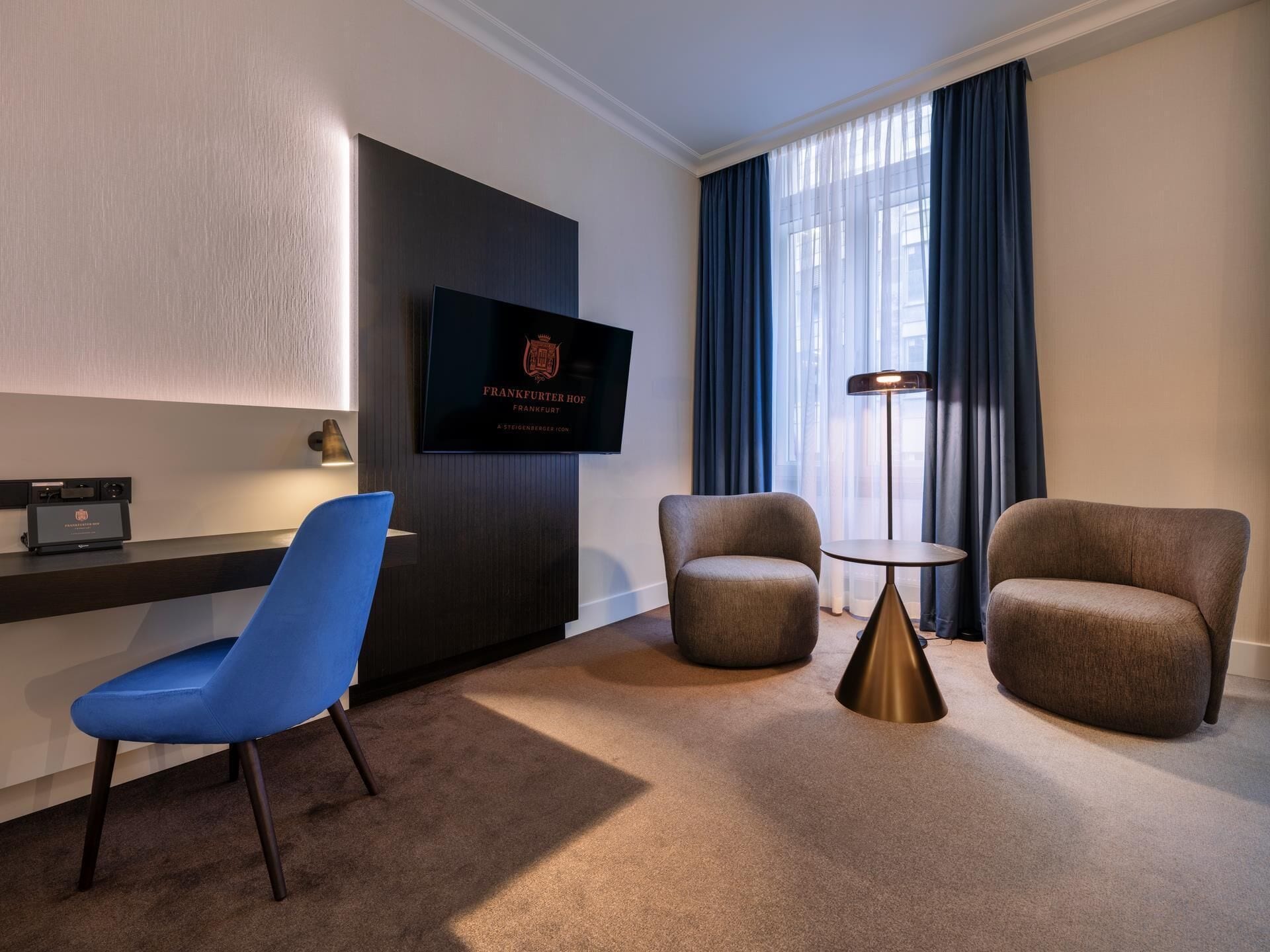 Ein Hotelzimmer mit flachbildfernseher an der Wand, zwei Sesseln, einem kleinen runden Tisch und einem blauen Sessel.