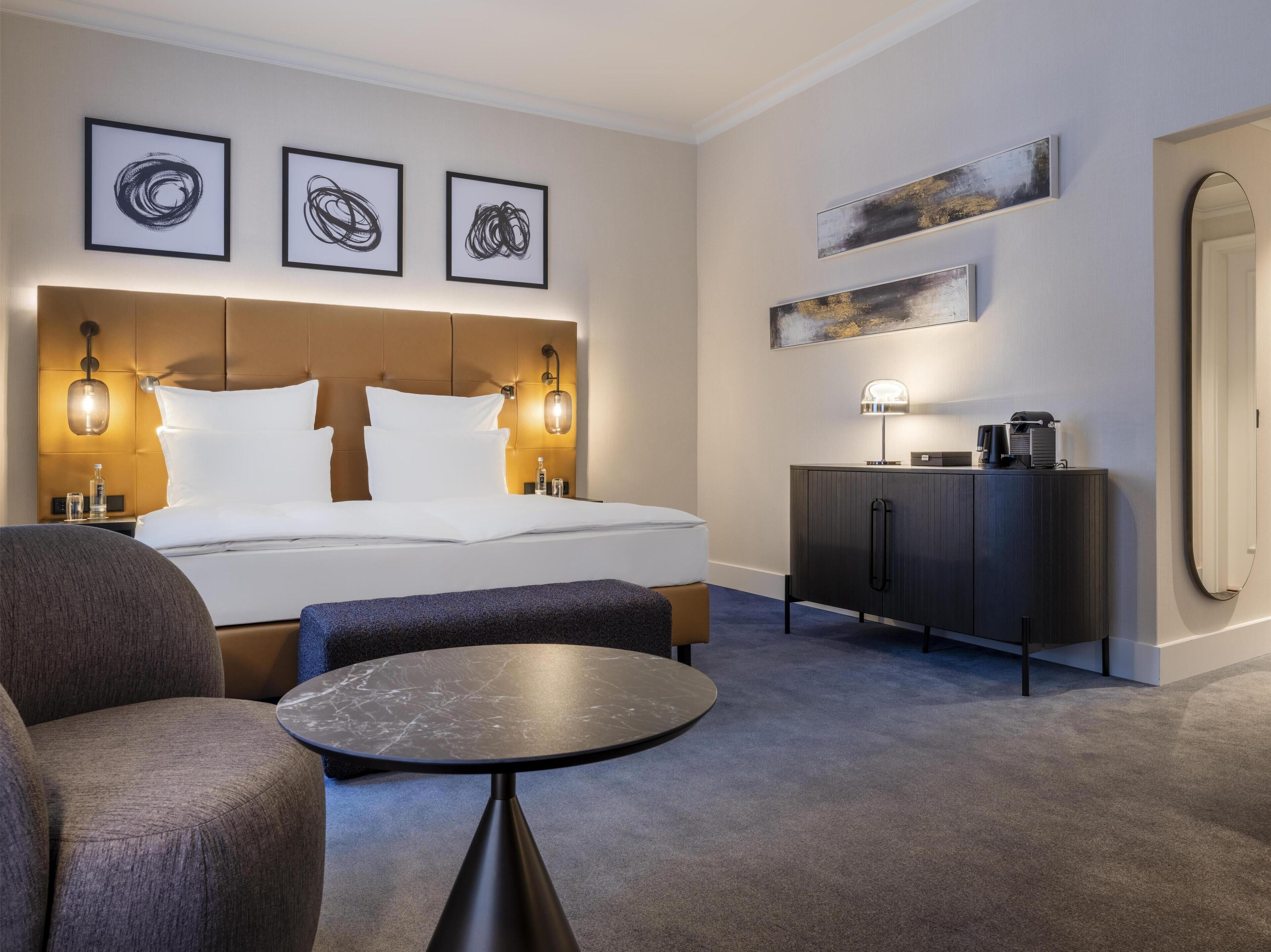 A hotel room with a large bed, a bedside table, a chair, a small round table, a cabinet, and artwork on the walls.