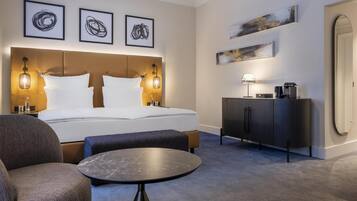 A hotel room with a large bed, a bedside table, a chair, a small round table, a cabinet, and artwork on the walls.