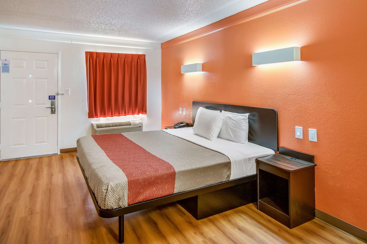 A hotel room with a bed, a bedside table, a nightstand, an air conditioning unit, and a window with curtains.