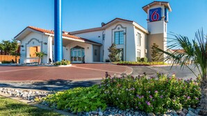 A hotel with a prominent '6' sign, a red-tiled roof, and a well-maintained garden with flowers and rocks. - Motel 6 Euless, TX - DFW West (Euless)