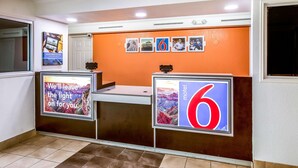 A hotel reception area with a reception desk, a large illuminated sign displaying the number 6, and a series of framed pictures on the wall. - Motel 6 Euless, TX - DFW West (Euless)