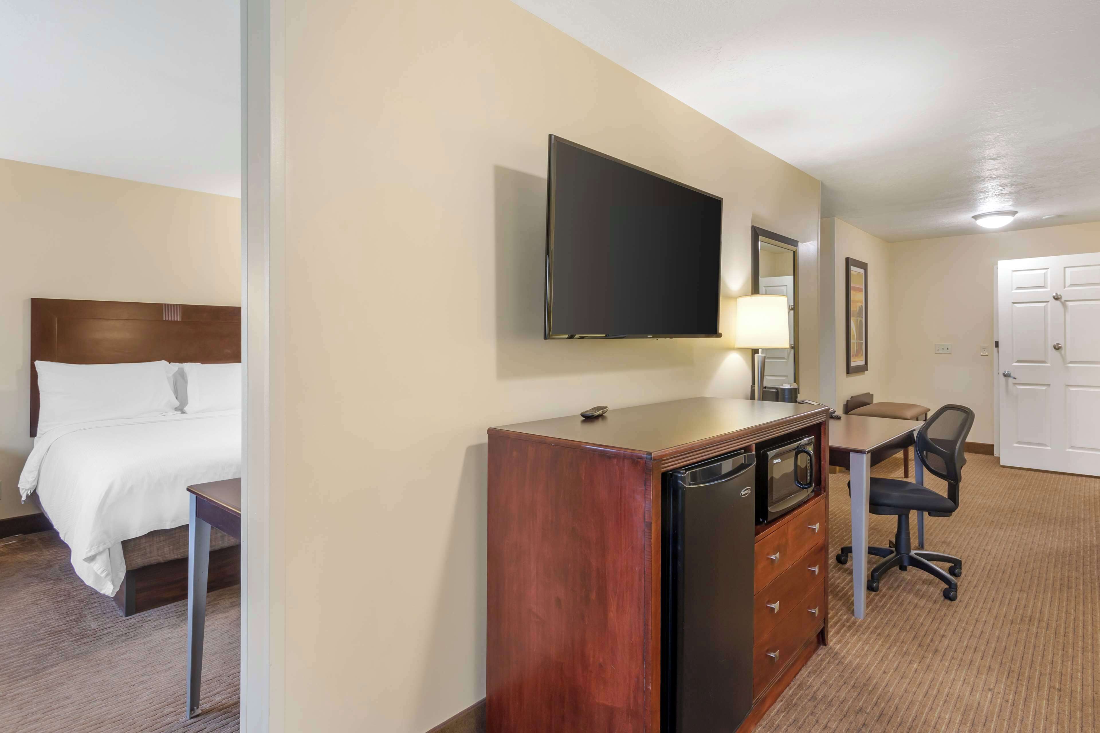 A hotel room with a bed, a desk with a chair, a television, a refrigerator, and a dresser.