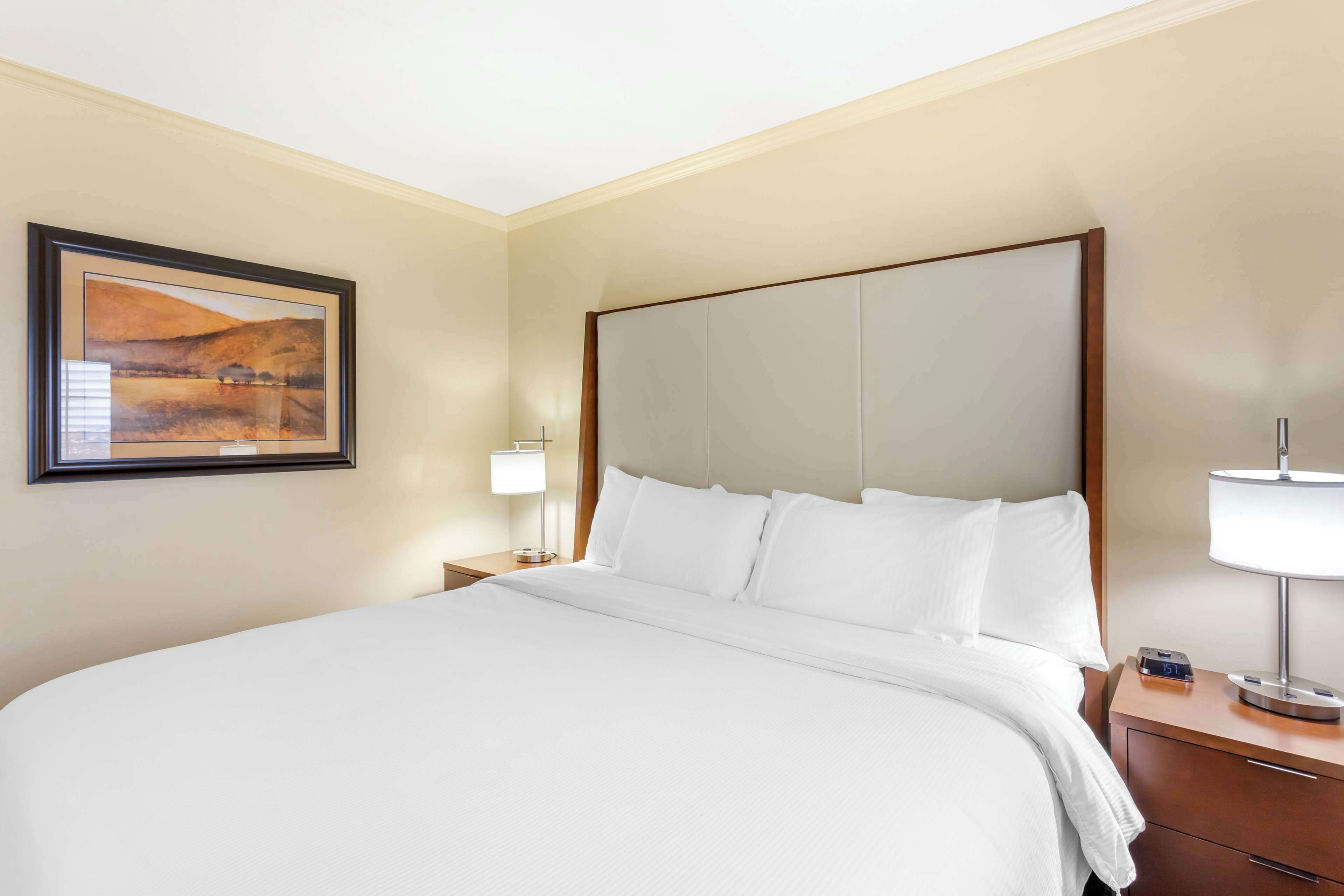 Una cama bien hecha con sábanas blancas, cabecera, mesita de noche con lámpara y un cuadro en la pared.