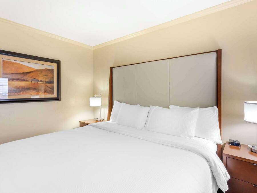 Una cama bien hecha con sábanas blancas, cabecera, mesita de noche con lámpara y un cuadro en la pared.