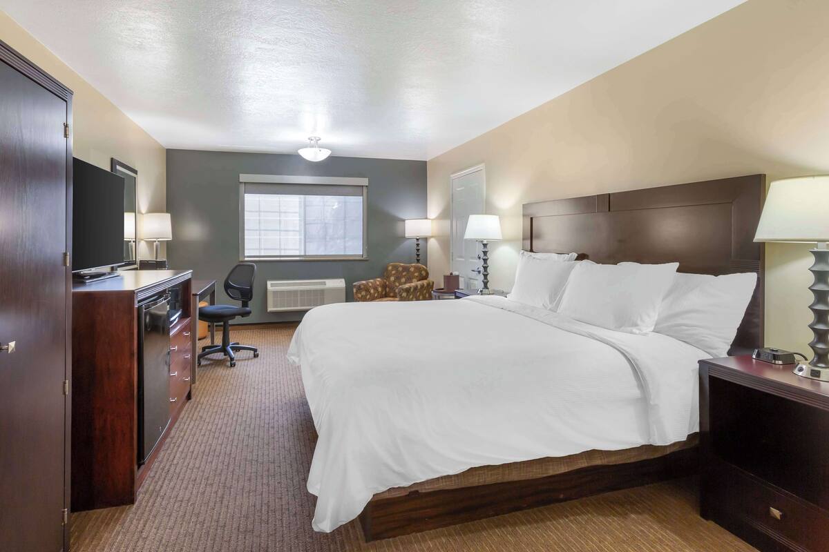 Une chambre d’hôtel avec un grand lit, un bureau avec une chaise, une armoire et une fenêtre avec des stores.