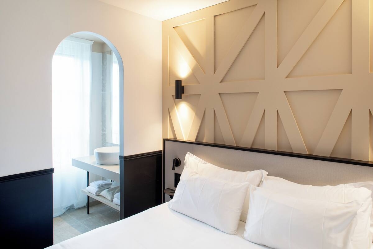 A modern hotel room with a large bed, a bedside table, a built-in shelf, a mirror, and a unique geometric wall design.