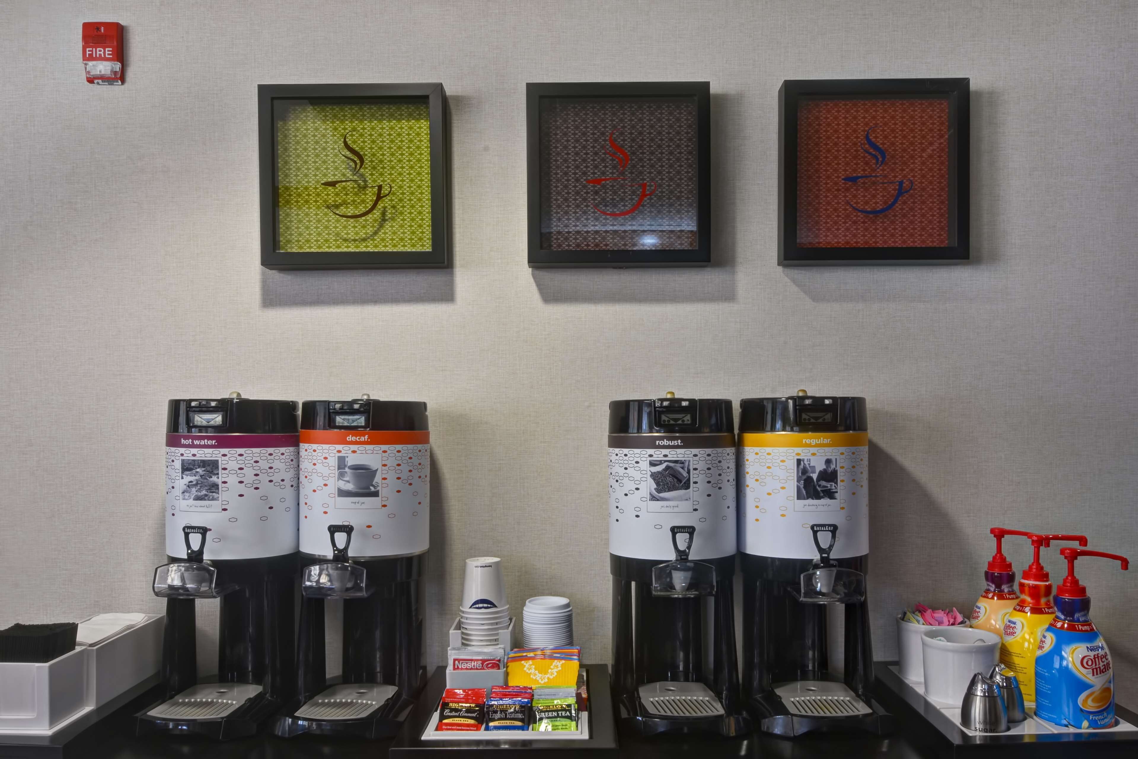 A coffee station with multiple dispensers, a fire extinguisher, and framed calligraphy art on the wall.