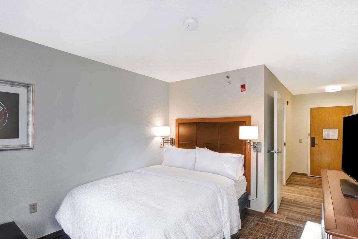 A hotel room with a large bed, a wooden headboard, a desk with a chair, a television, and a door leading to another room.