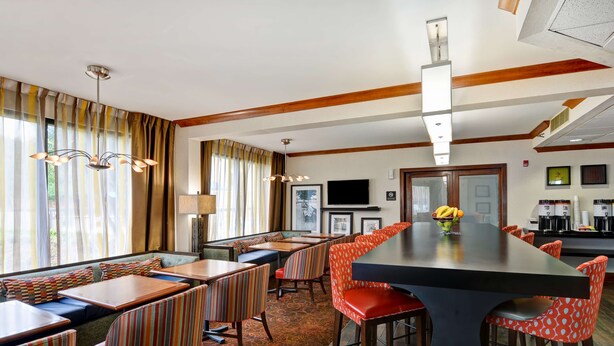 A hotel dining area with striped booths, a long communal table, and a kitchen in the background. - Hampton Inn Memphis-Poplar (Memphis)