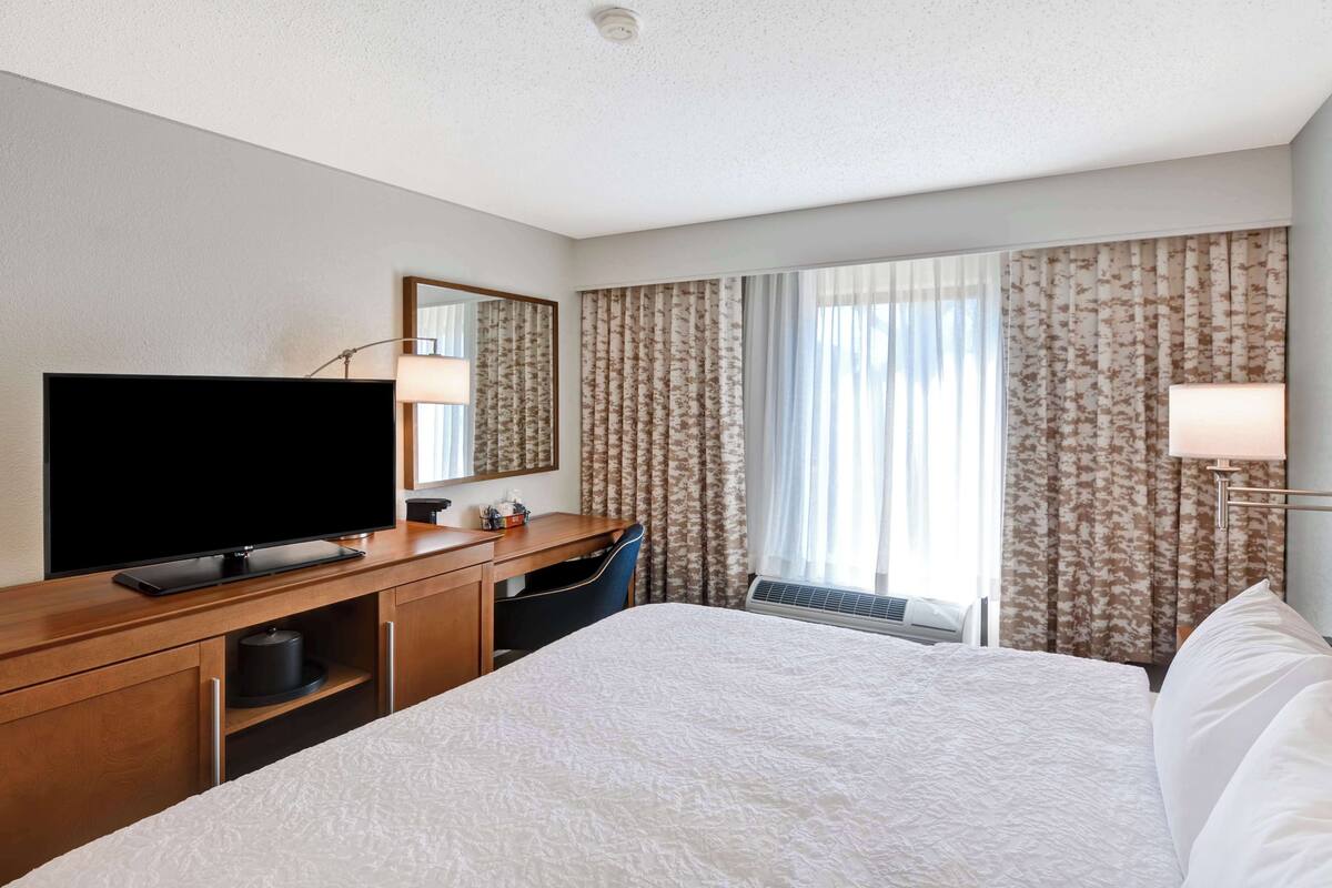 A hotel room with a large bed, a flat-screen TV, a desk with a chair, and a window with curtains.