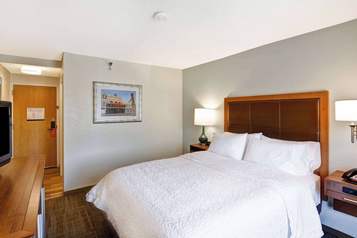 A hotel room with a large bed, a wooden headboard, a desk with a chair, a television, and a framed picture on the wall.