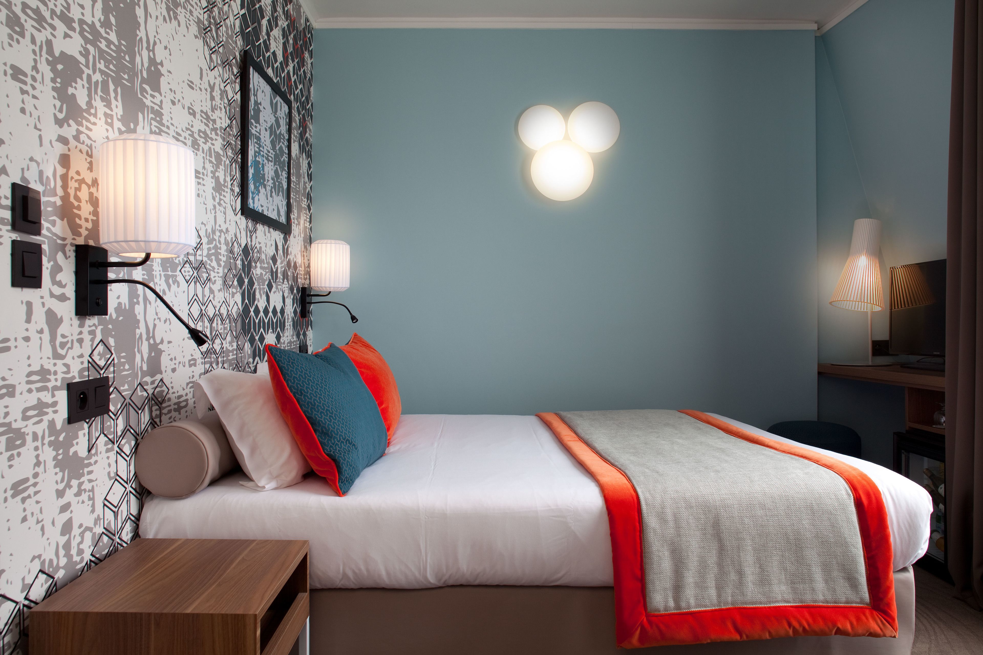 A modern hotel room with a neatly made bed, a bedside table, and a desk with a lamp.