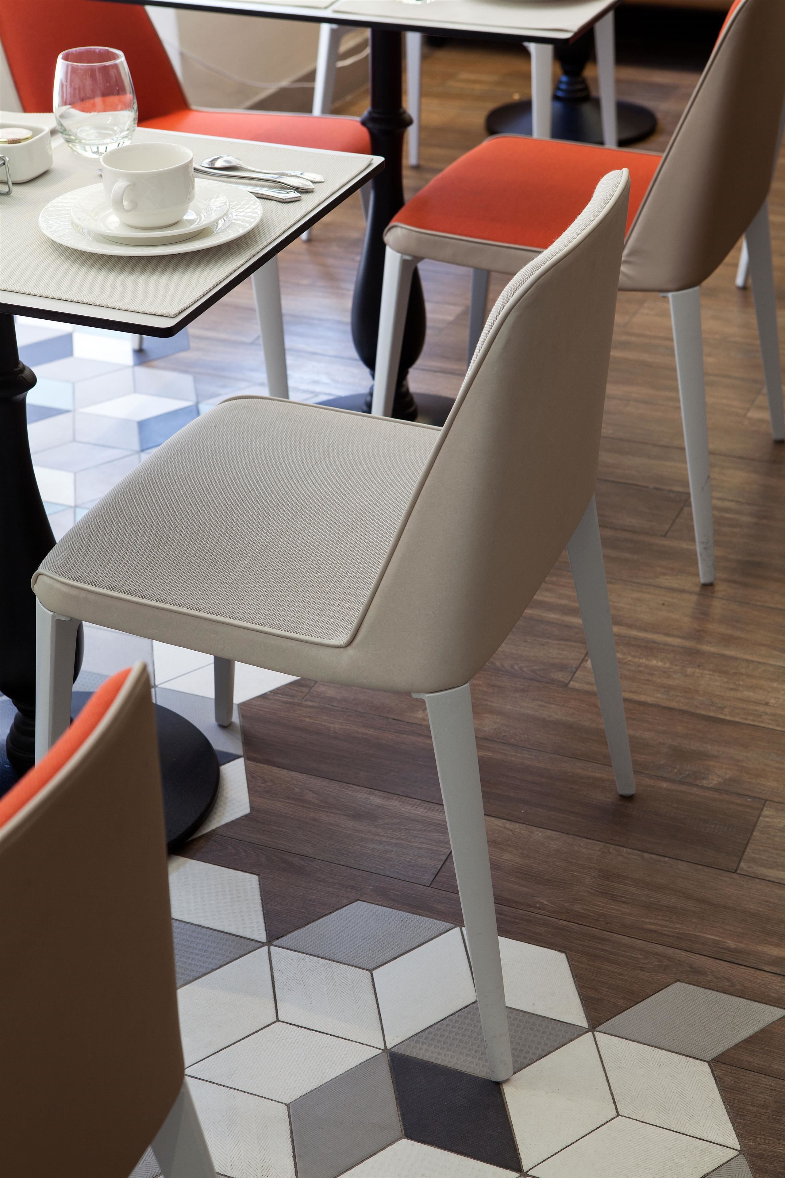 Een eetgedeelte met een tafel gedekt voor twee personen, bestaande uit een witte kop en schotel, een glas water en bestek. De stoelen hebben oranje kussens, en de vloer heeft een geometrisch tegelpatroon.