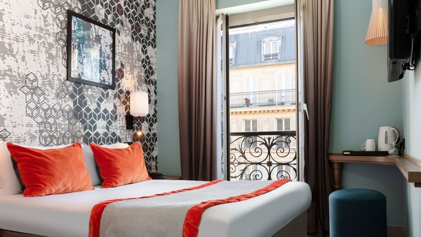 A hotel room with a bed, a view of the city, a small table with a kettle, and a TV mounted on the wall. - Hotel Des Nations Saint Germain (Paris)