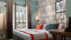 A hotel room with a large bed, a bedside table, a lamp, and a view of the city through a window. - Hotel Des Nations Saint Germain (Paris)