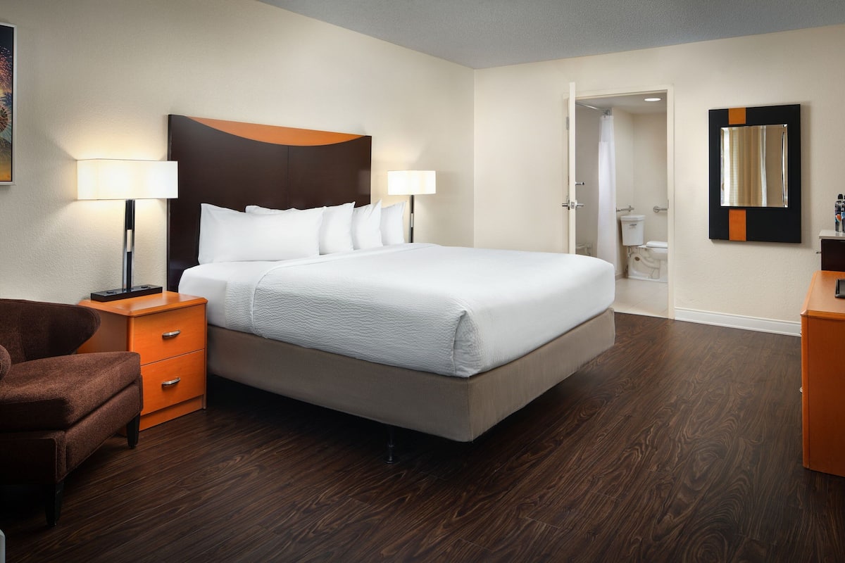 Una habitación de hotel con una cama grande, dos lámparas de noche, una mesita de noche de madera, un sillón marrón y un aparador de madera.