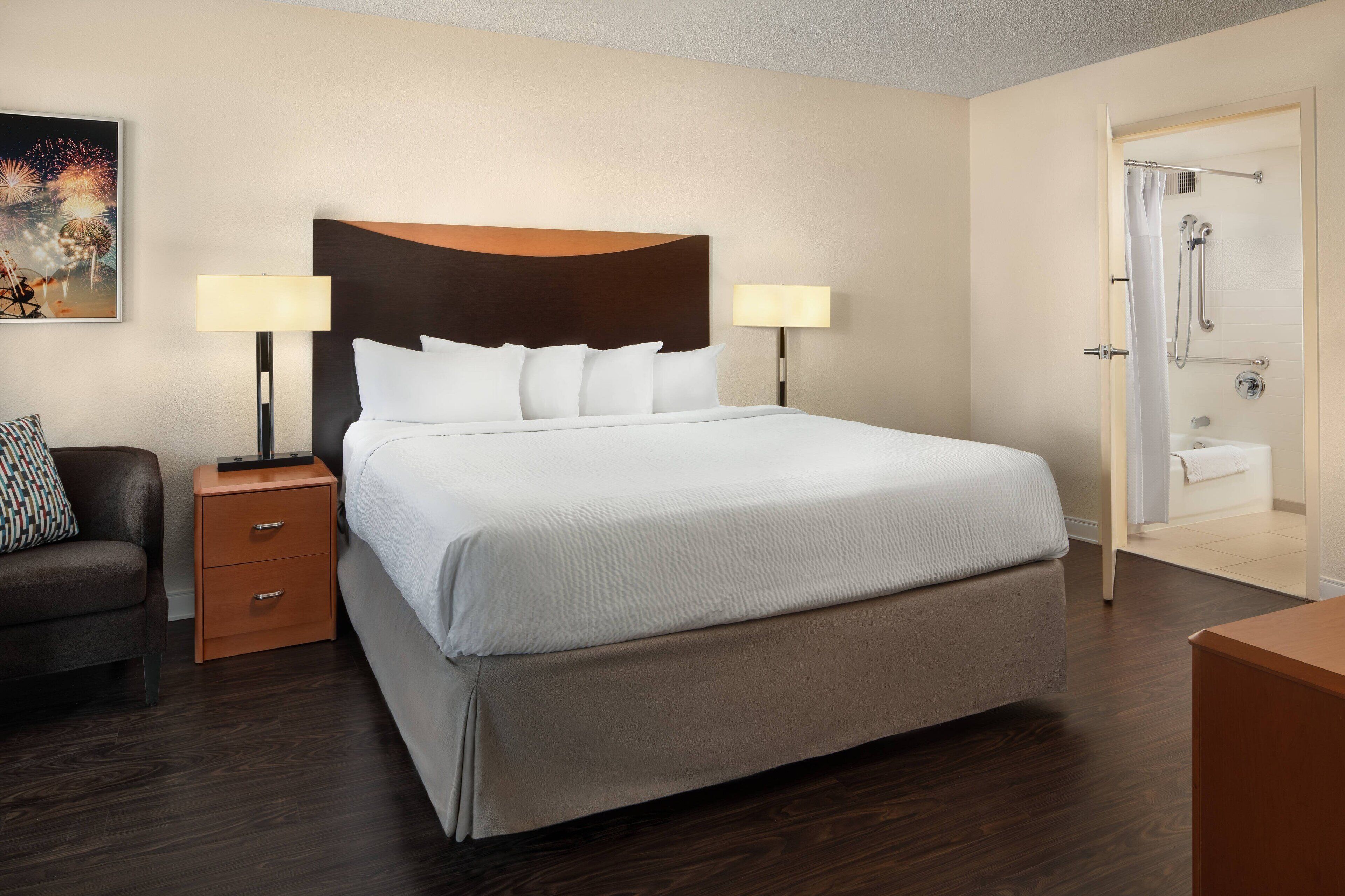 A hotel room with a large bed, two bedside tables with lamps, a dark wood floor, a shower area, and a painting on the wall.