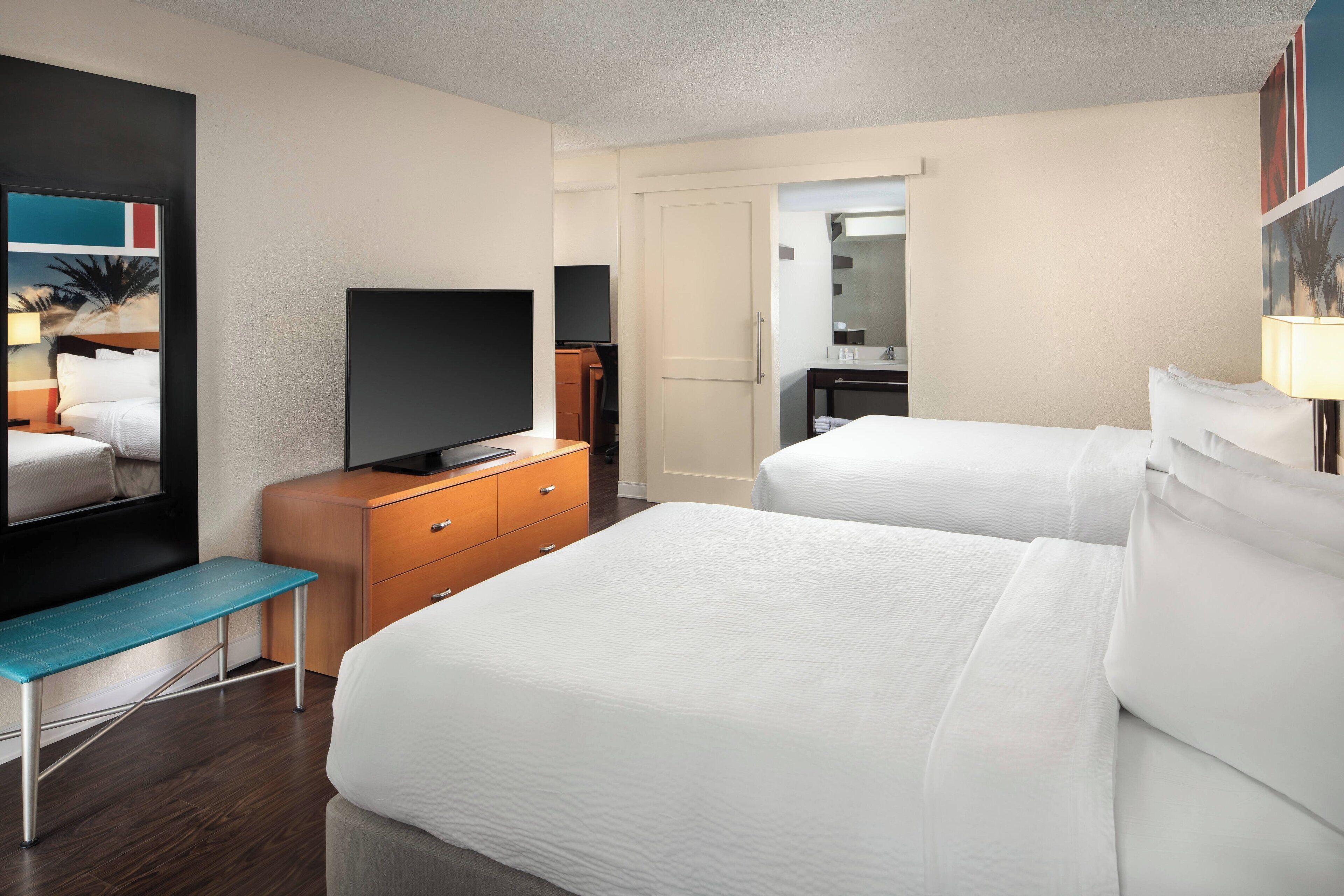 Habitación de hotel con dos camas, un televisor sobre una cómoda de madera, un banco y un baño visible a través de una puerta abierta.