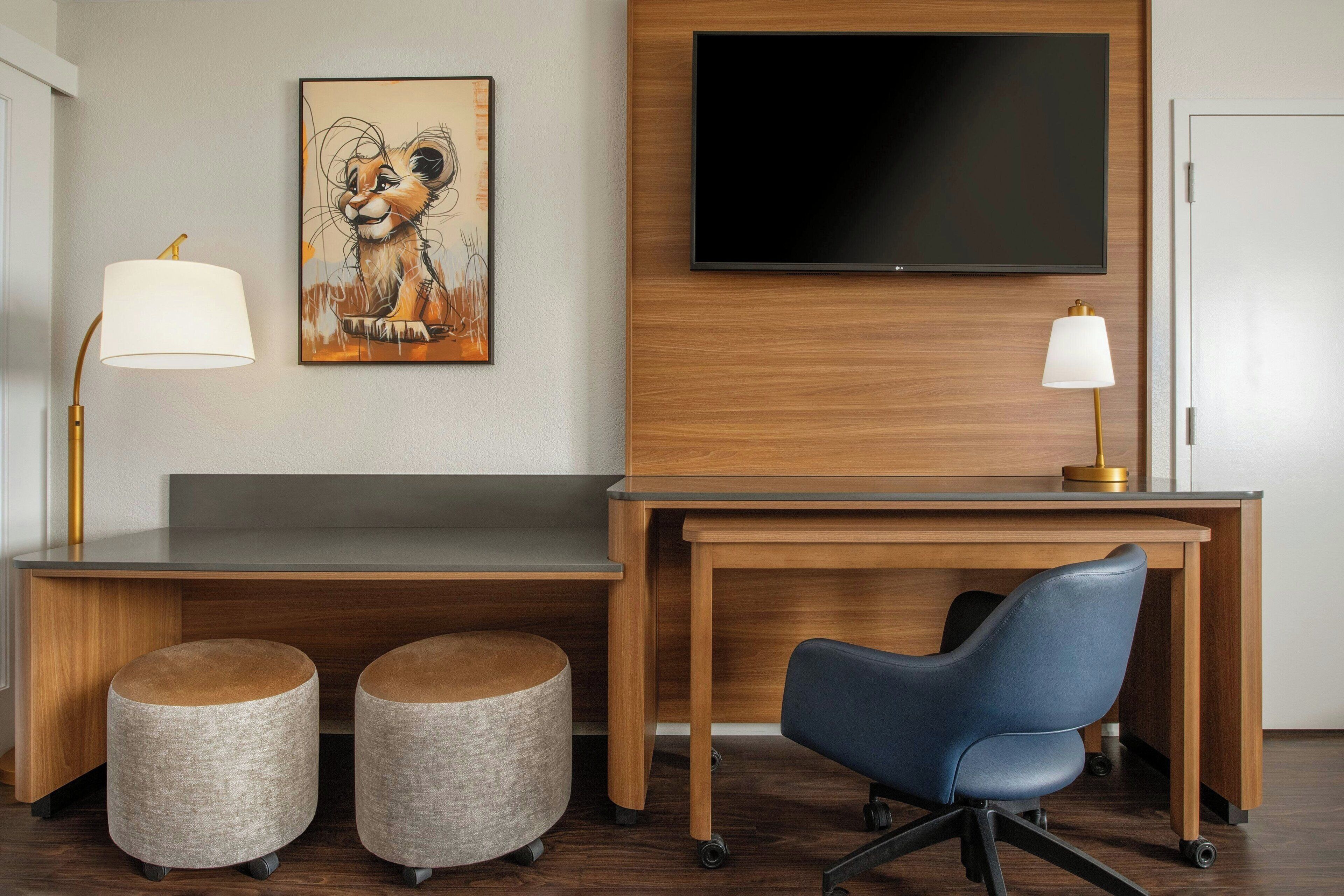A hotel room with a wooden desk, two ottomans, a blue office chair, a flat-screen TV, and two lamps.