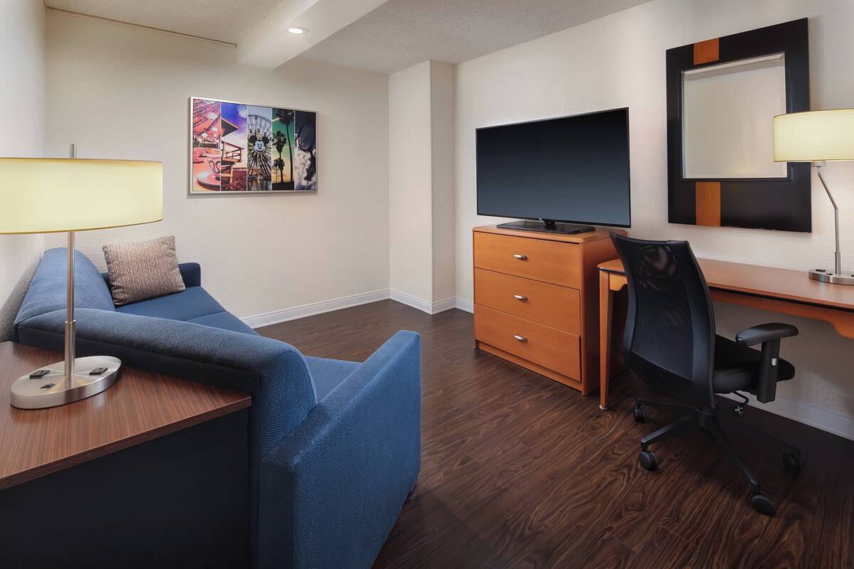 Una habitación de hotel moderna con sofá, escritorio con lámpara, televisión y cómoda de madera.