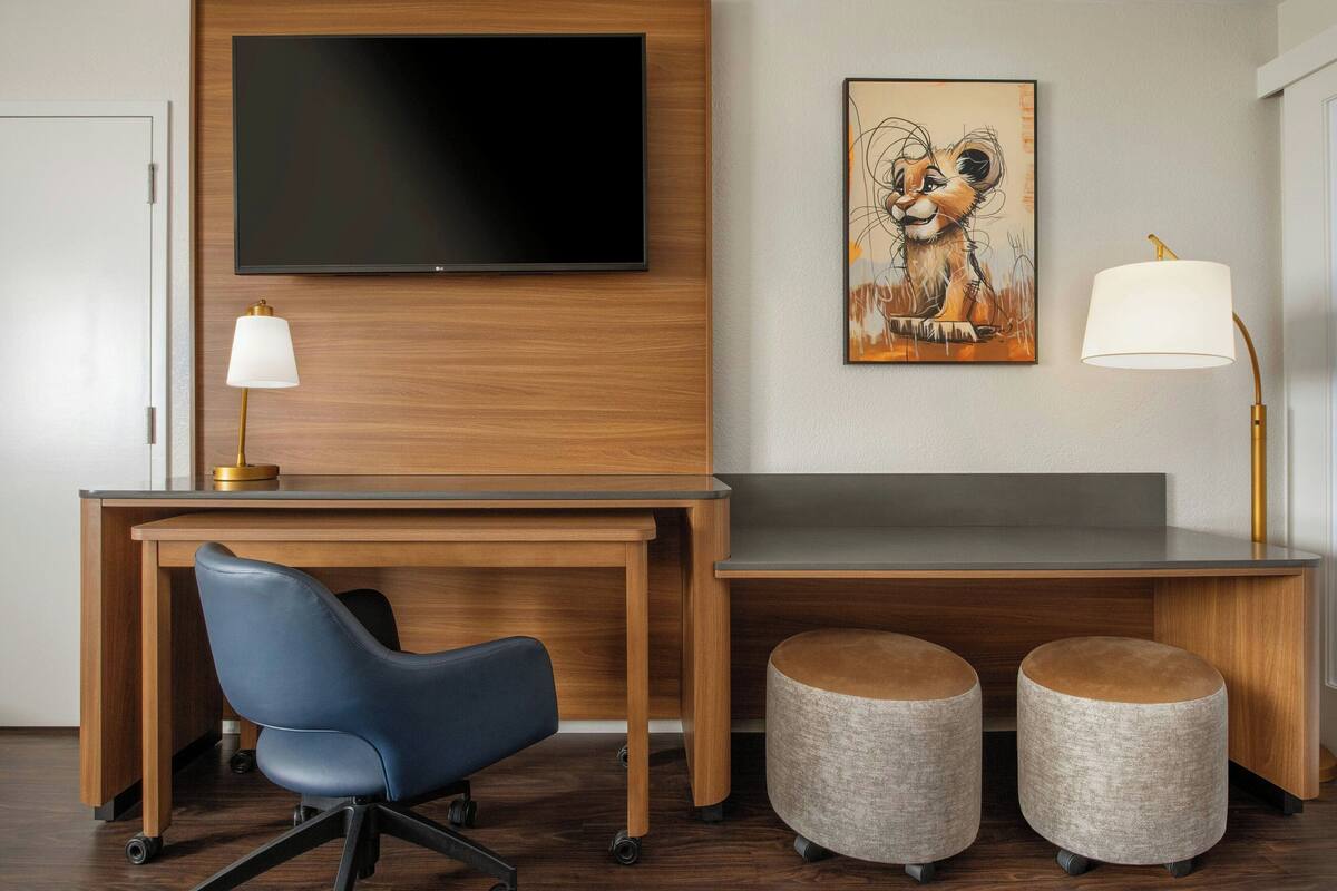 Habitación de hotel moderna con un escritorio de madera, una silla de oficina azul, dos otomanas, un televisor de pantalla plana, una lámpara de pared y una obra de arte enmarcada.