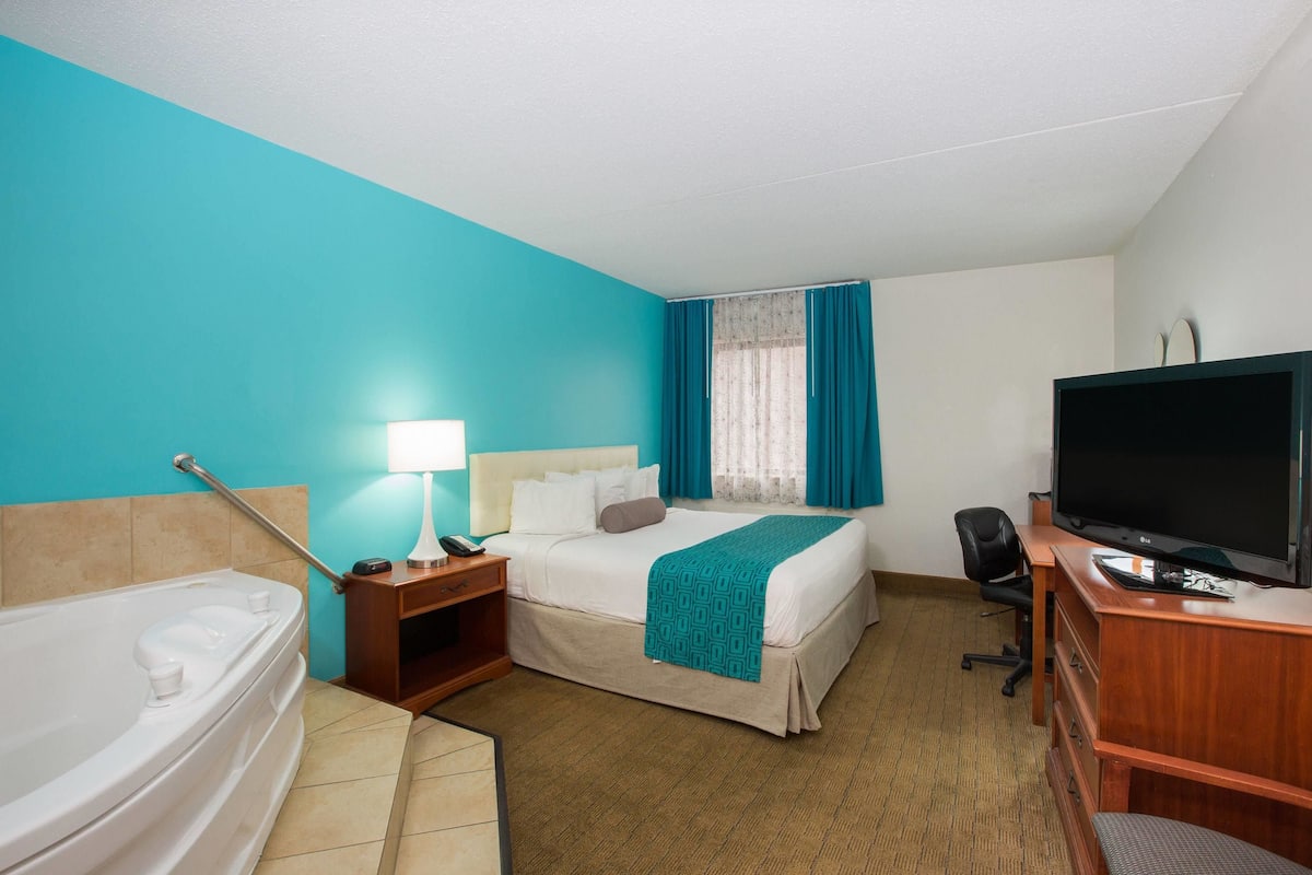 A hotel room with a bed, a desk with a television, a desk lamp, a chair, a nightstand, and a bathtub.