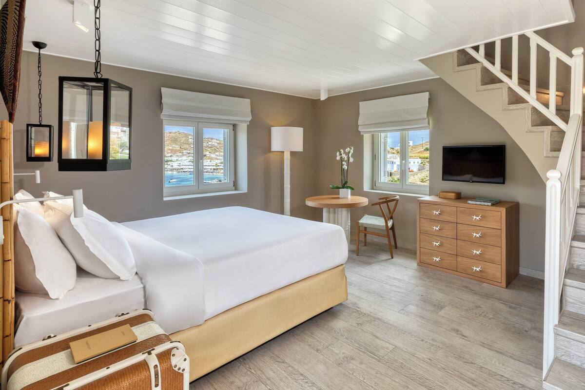 A bedroom with a large bed, a desk, a chair, a TV, and a staircase.